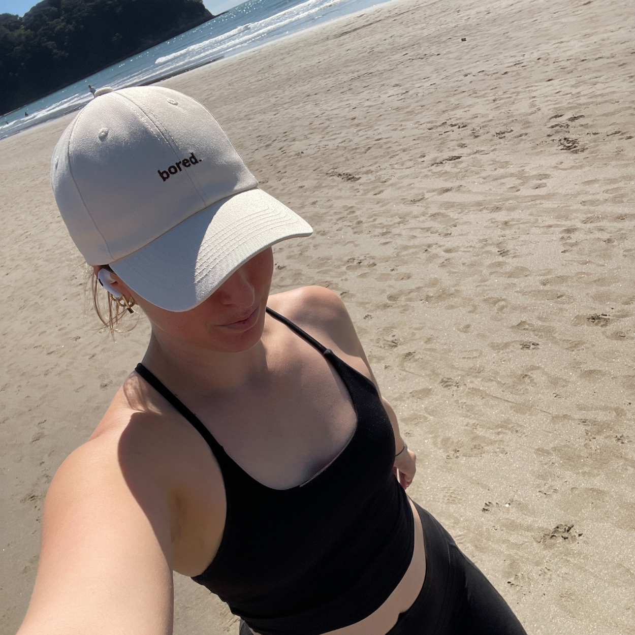 Woman on the beach wearing beige baseball cap with "bored" embroidered on the front