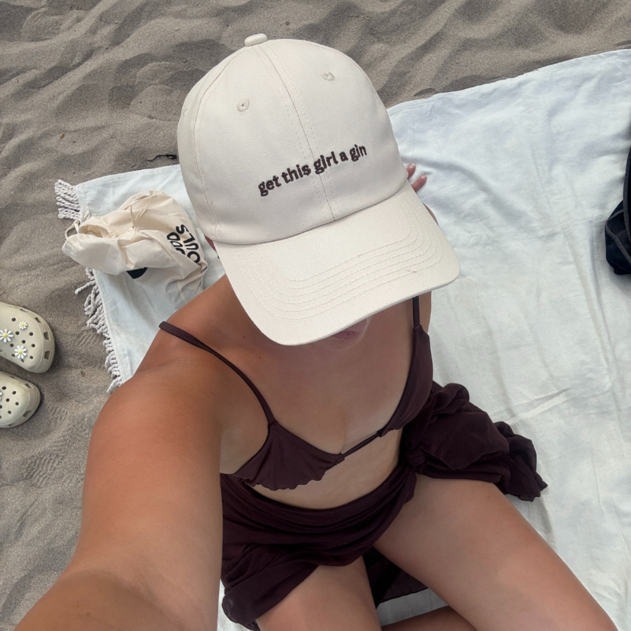 Woman on the beach wearing beige embroidered cap that says "get this girl a gin"