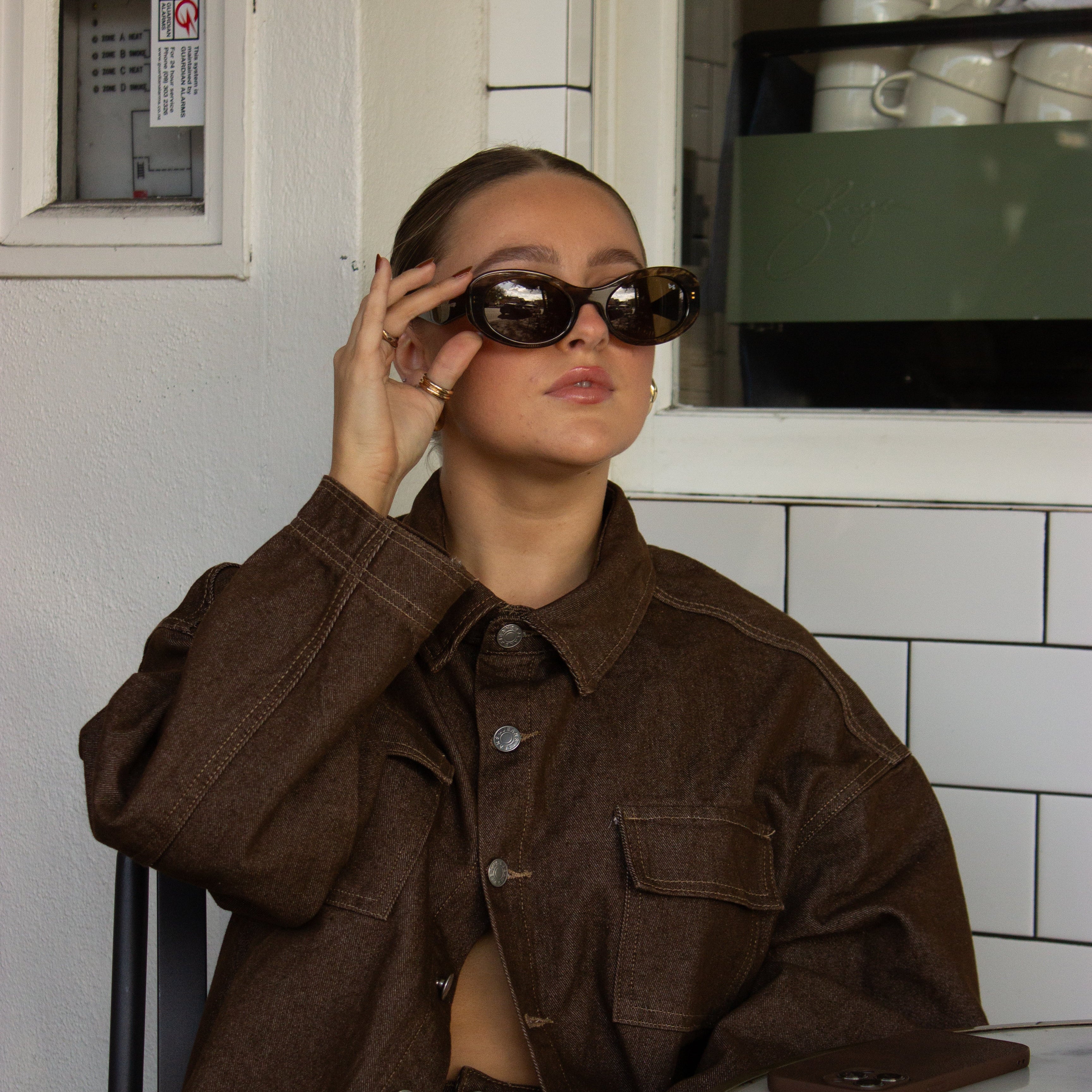 Woman wearing polarised chocolate espresso oval sunglasses and a brown jacket in an indoor setting